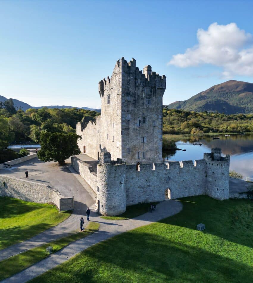 Stone castle with a central tower stands beside a lake, surrounded by green grass and trees, with mountains and blue sky in the background.
