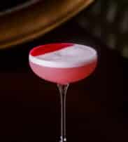 A pink cocktail with a foamy white top and a semicircle of red garnish, served in a coupe glass against a dark background.