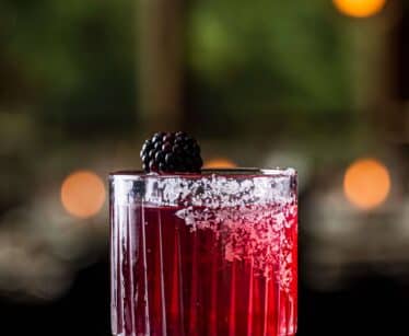 A glass of red cocktail with a salted rim, garnished with a blackberry, sits on a reflective surface with blurred lights in the background.