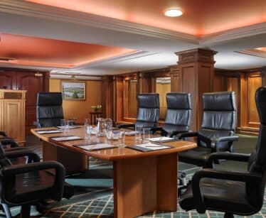 A conference room with a wooden table, six black high-backed chairs, notepads, water glasses, a jug, and a lectern in the background.