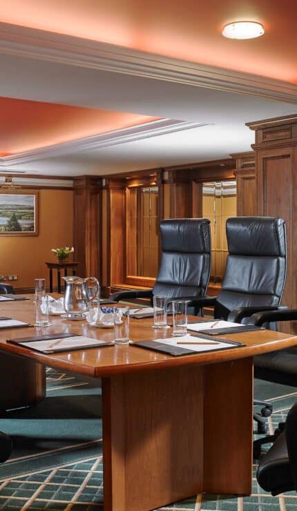 A conference room with a wooden table, six black high-backed chairs, notepads, water glasses, a jug, and a lectern in the background.