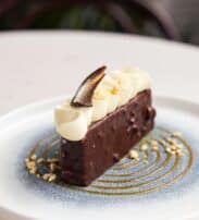 A slice of chocolate-covered cake topped with white cream swirls, a chocolate shard, and chopped nuts, on a blue and white plate with a decorative swirl and nut pieces.