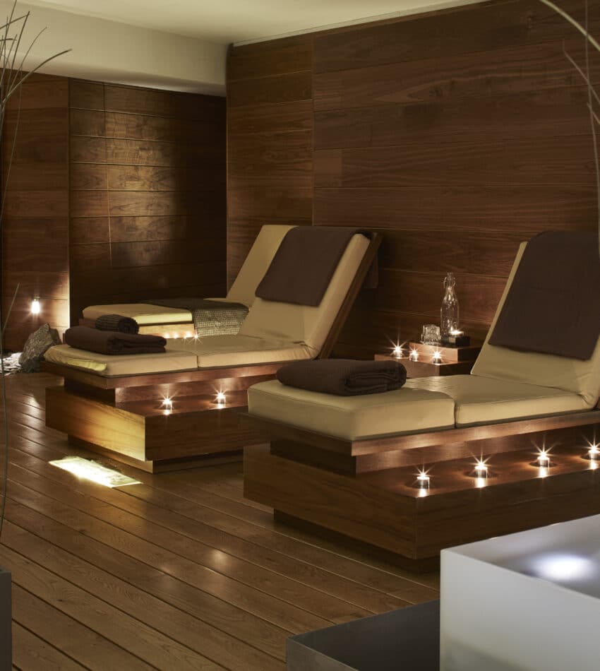 Two lounge chairs with brown cushions and towels are set in a modern spa room with wooden walls, soft lighting, and decorative plants in the foreground.