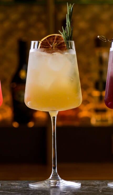 Three cocktails in wine glasses with ice, each garnished differently: one with a strawberry and mint, one with dried citrus and rosemary, and one with blackberries and a twig.