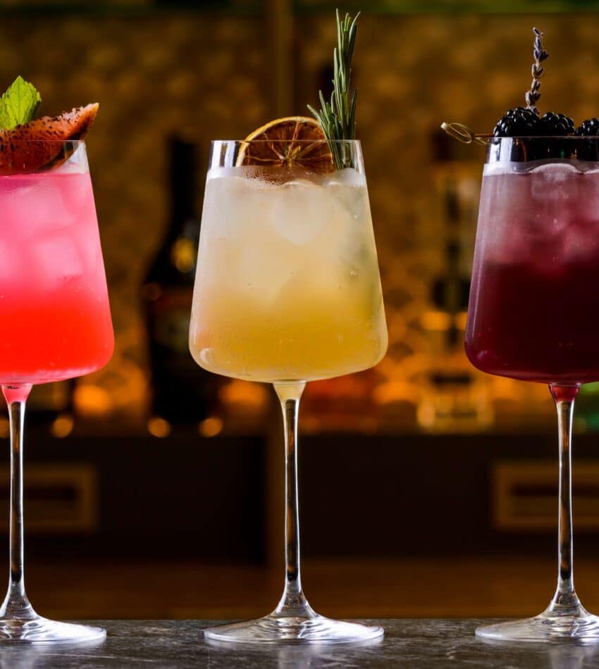 Three cocktails in wine glasses with ice, each garnished differently: one with a strawberry and mint, one with dried citrus and rosemary, and one with blackberries and a twig.