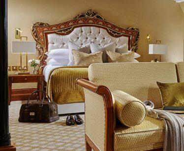 Elegant hotel room with a large, ornate bed, gold and white bedding, matching furniture, a brown travel bag, and shoes on a patterned carpet.