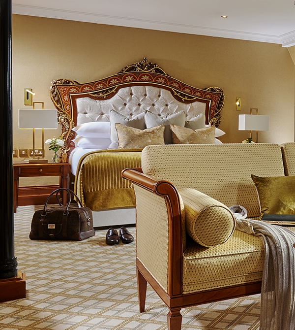 Elegant hotel room with a large, ornate bed, gold and white bedding, matching furniture, a brown travel bag, and shoes on a patterned carpet.