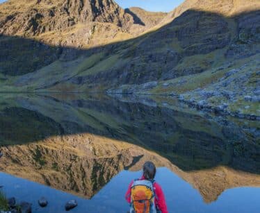 A person with an orange rucksack stands by a calm mountain lake, looking at the reflected peaks and clear blue sky.