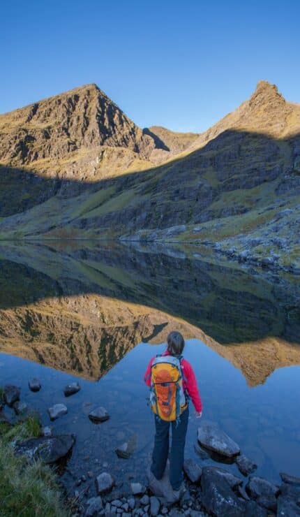 A person with an orange rucksack stands by a calm mountain lake, looking at the reflected peaks and clear blue sky.