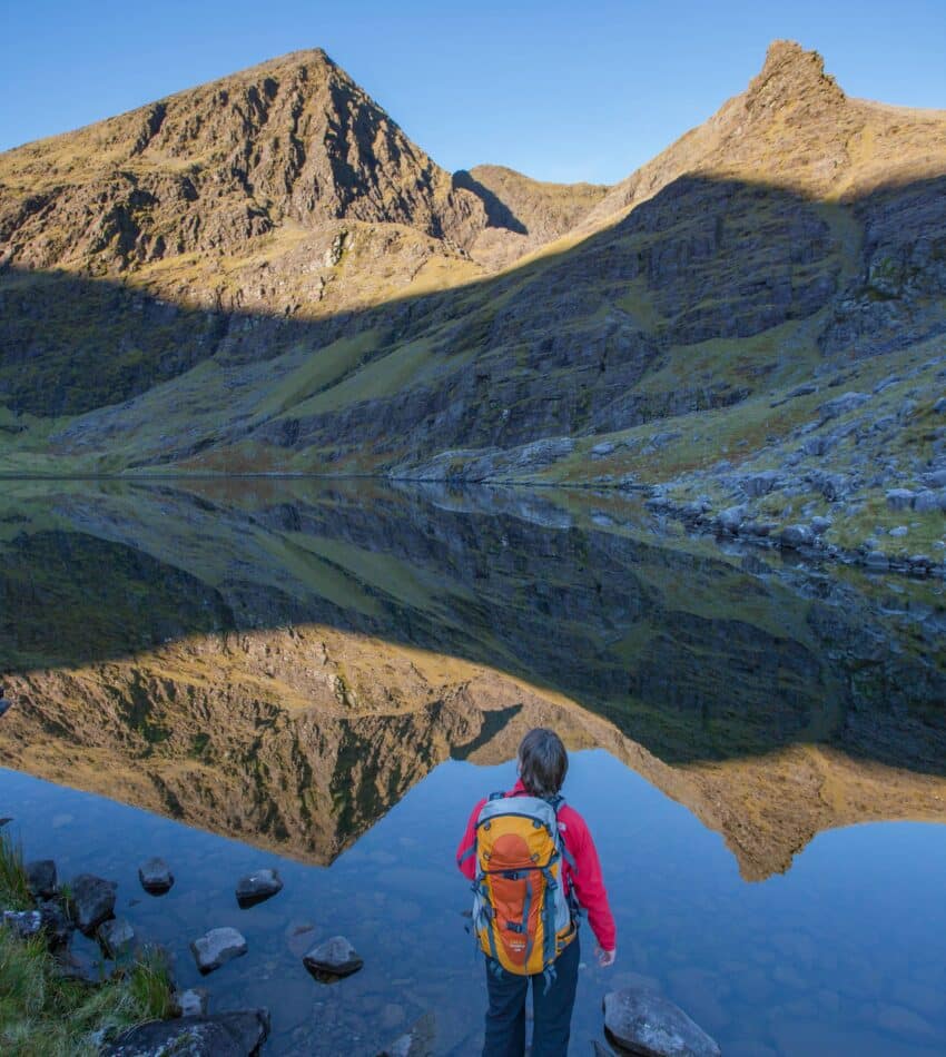 A person with an orange rucksack stands by a calm mountain lake, looking at the reflected peaks and clear blue sky.