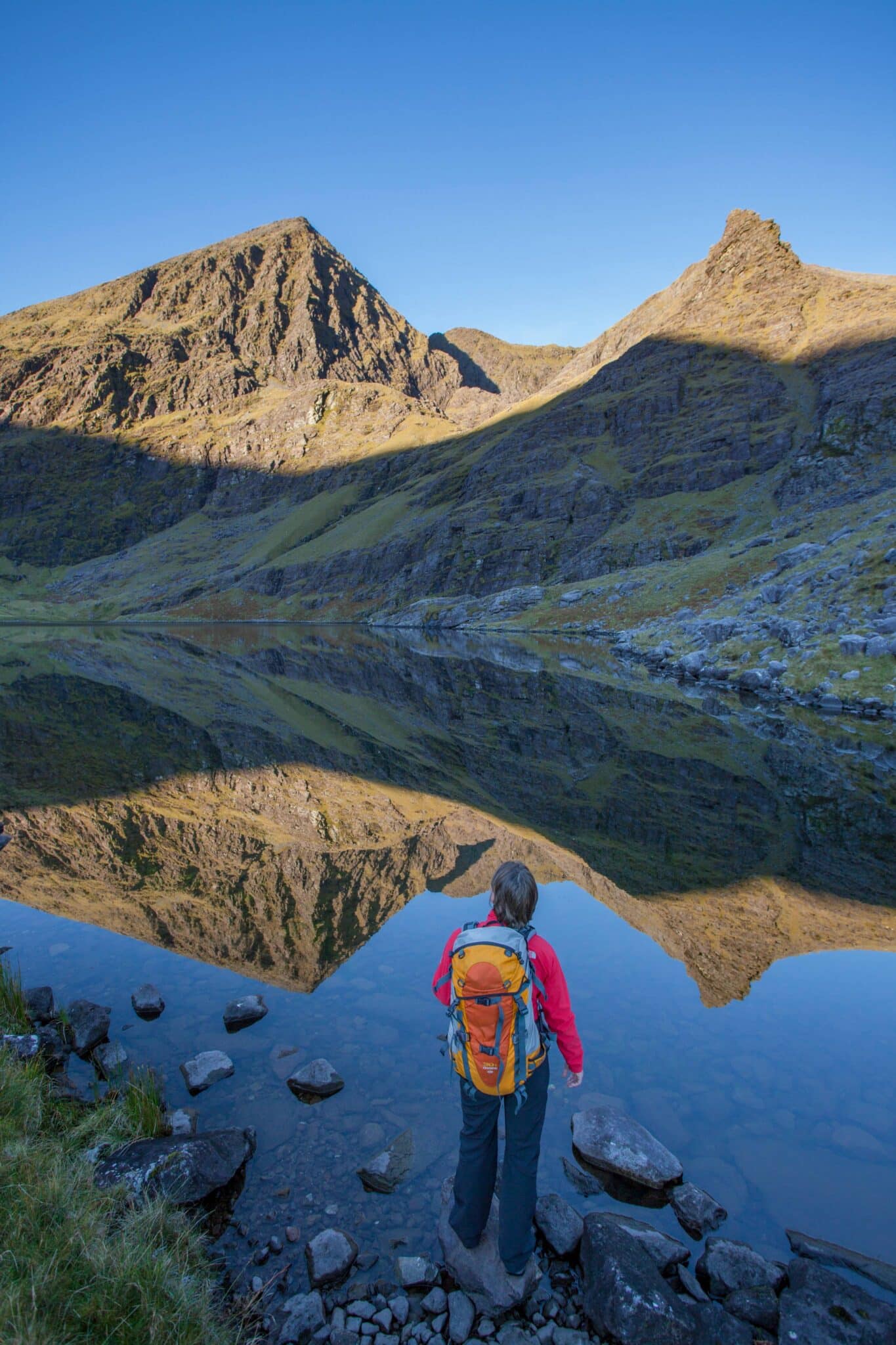 A person with an orange rucksack stands by a calm mountain lake, looking at the reflected peaks and clear blue sky.