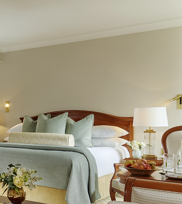 A neatly made bed with multiple pillows and a throw blanket, a bedside table with a lamp and flowers, and a tray with fruit and water on a table in a hotel room.