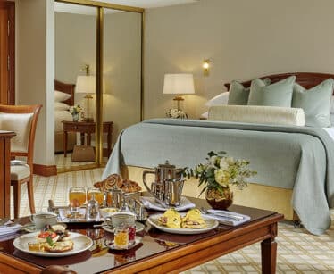 A hotel room with a made bed, mirrored wardrobe, and a table set with breakfast items, pastries, coffee, and fresh flowers.