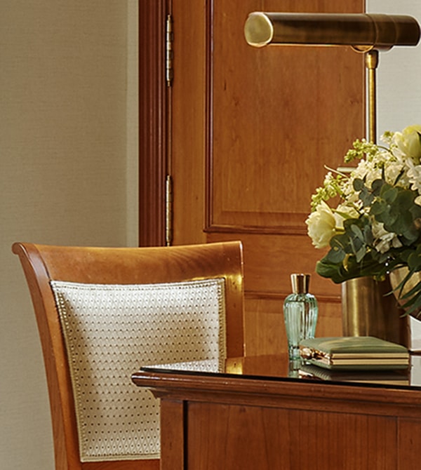 A wooden chair and desk with a gold lamp, a vase of white flowers, a blue glass bottle, and books, set against a wooden door.