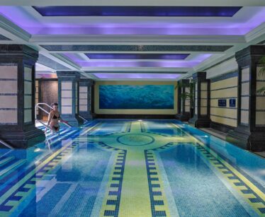 Indoor swimming pool with decorative tiles, blue lighting, and a woman sitting on the edge of the pool near metal handrails.
