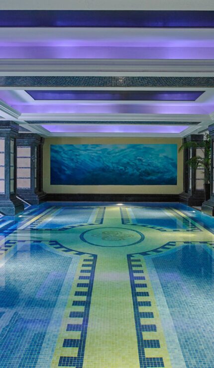 Indoor swimming pool with decorative tiles, blue lighting, and a woman sitting on the edge of the pool near metal handrails.