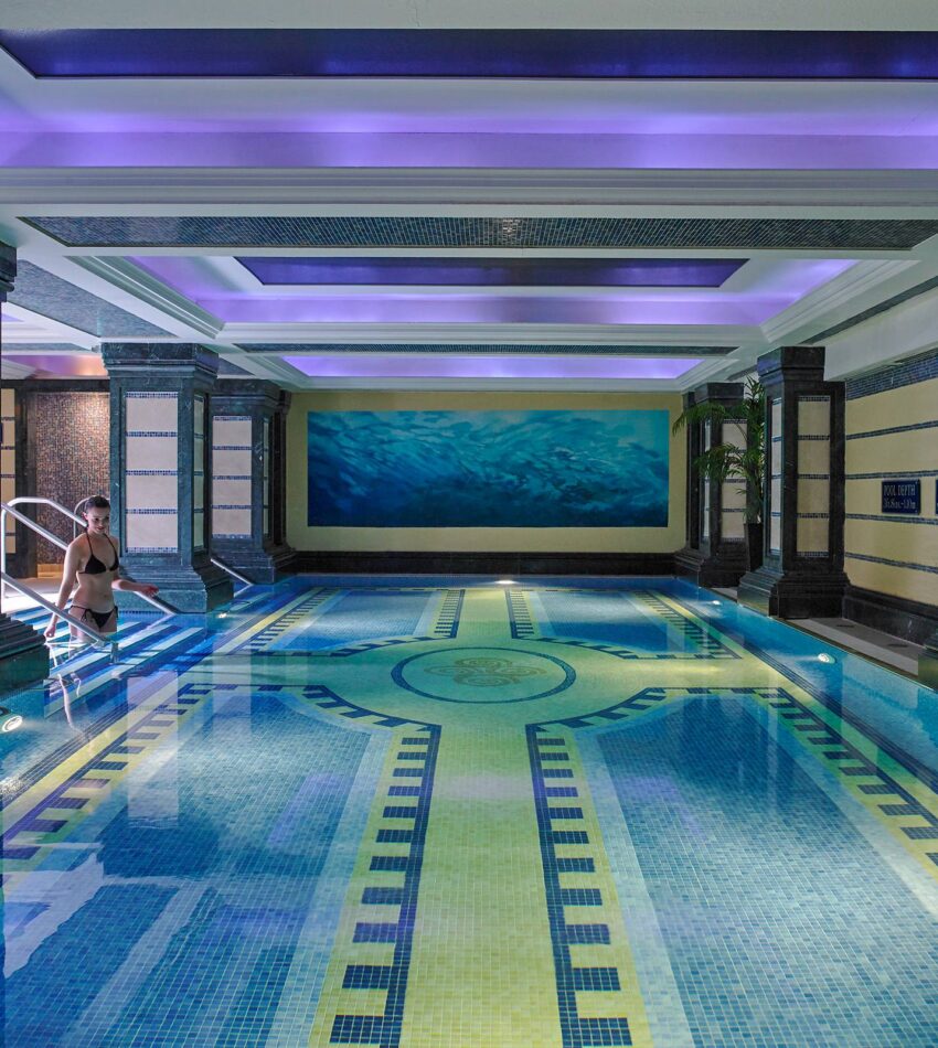 Indoor swimming pool with decorative tiles, blue lighting, and a woman sitting on the edge of the pool near metal handrails.