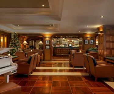 A hotel lounge with brown leather chairs, a white piano, a decorated Christmas tree, and a fully stocked bar in the background.