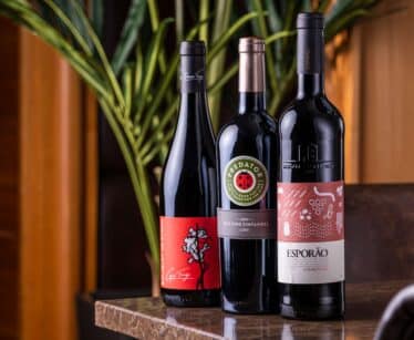 Three bottles of red wine are displayed upright on a marble surface with green leaves and wooden panelling in the background.