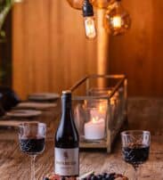 A bottle of Paparuda Pinot Noir wine with two filled glasses and a platter of cheese, nuts, and grapes on a wooden table, lit by hanging bulbs and a candle.