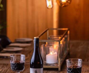 A bottle of Paparuda Pinot Noir wine with two filled glasses and a platter of cheese, nuts, and grapes on a wooden table, lit by hanging bulbs and a candle.