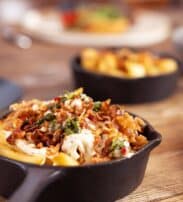 A small cast iron frying pan filled with chips topped with melted cheese, bacon pieces, and herbs sits on a wooden table. Another frying pan with a similar dish is blurred in the background.