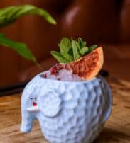 A blue elephant-shaped mug filled with crushed ice, garnished with a dried orange slice, mint leaves, and red petals, sits on a wooden surface.