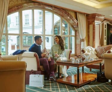 A man and woman sit on a sofa in a hotel lounge, smiling and holding drinks, with a shopping bag on the floor and large arched windows in the background.