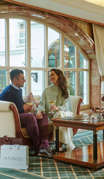 A man and woman sit on a sofa in a hotel lounge, smiling and holding drinks, with a shopping bag on the floor and large arched windows in the background.