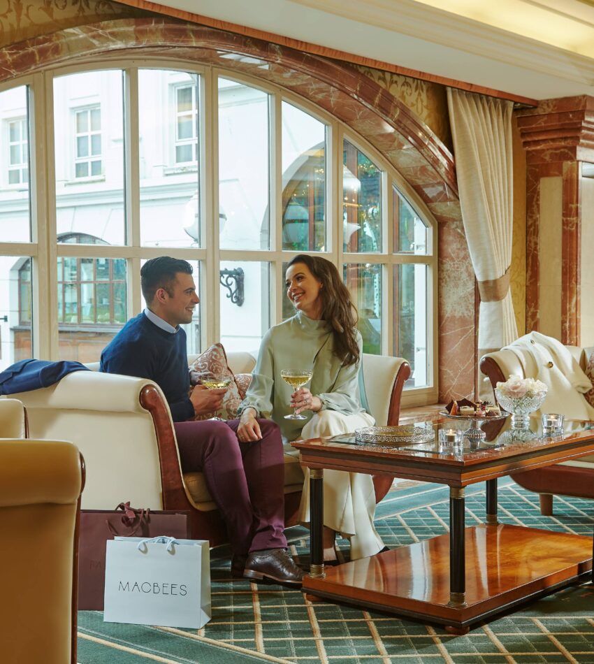 A man and woman sit on a sofa in a hotel lounge, smiling and holding drinks, with a shopping bag on the floor and large arched windows in the background.