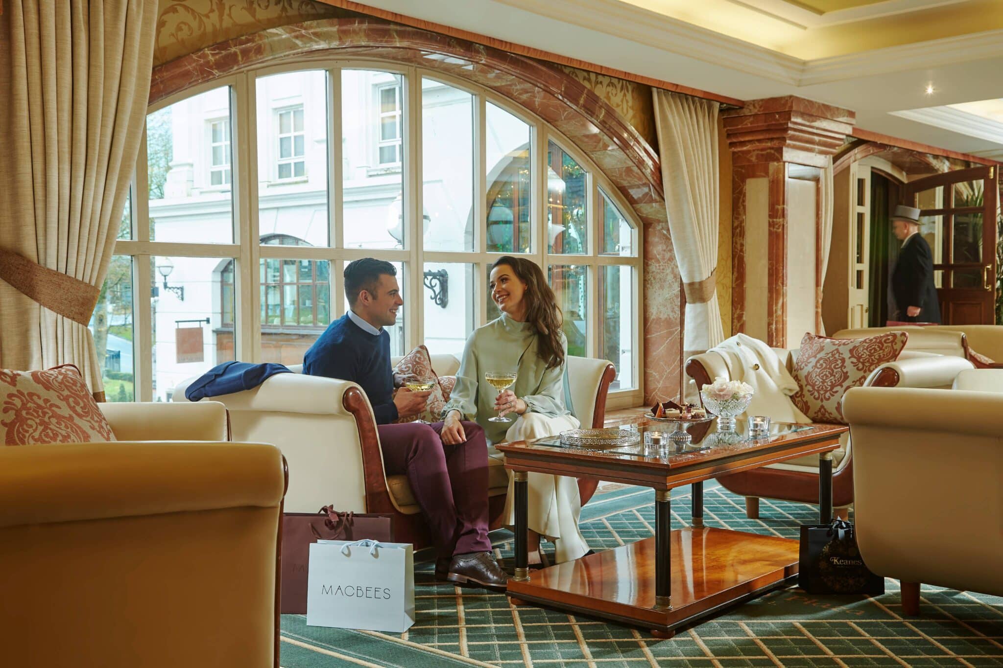 A man and woman sit on a sofa in a hotel lounge, smiling and holding drinks, with a shopping bag on the floor and large arched windows in the background.