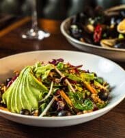 A bowl of mixed salad with sliced avocado, olives, and greens on a wooden table, beside a plate of mussels and a glass of white wine.