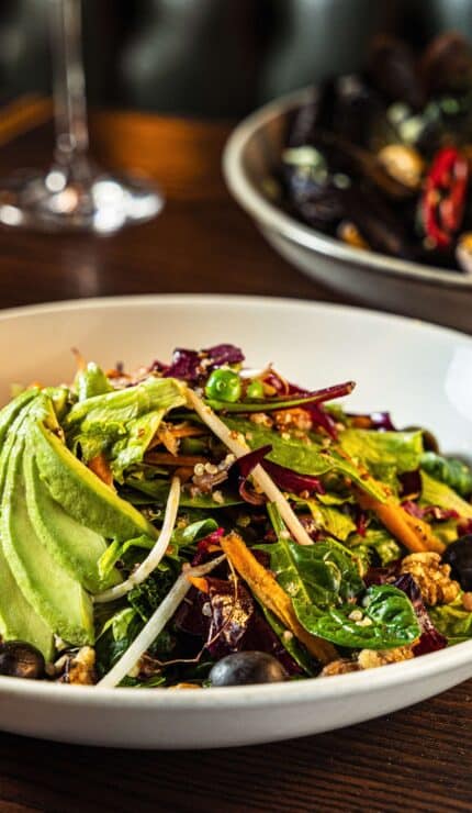 A bowl of mixed salad with sliced avocado, olives, and greens on a wooden table, beside a plate of mussels and a glass of white wine.