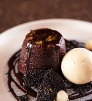 A chocolate lava pudding topped with chocolate sauce, served with a scoop of vanilla ice cream and garnished with black sesame tuile on a white plate.