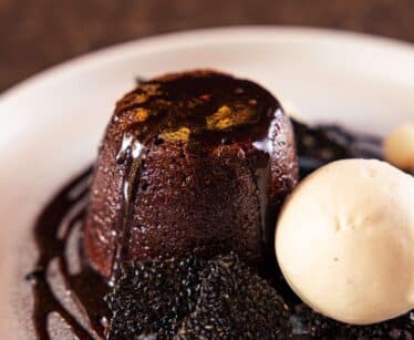 A chocolate lava pudding topped with chocolate sauce, served with a scoop of vanilla ice cream and garnished with black sesame tuile on a white plate.