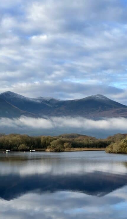 A calm lake reflects distant mountains, trees, and a cloudy sky with low-lying fog. Several small boats are scattered on the water.