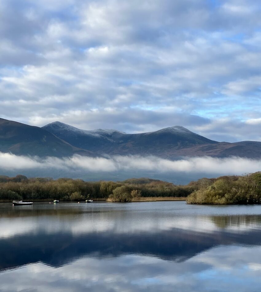 A calm lake reflects distant mountains, trees, and a cloudy sky with low-lying fog. Several small boats are scattered on the water.