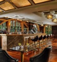 A barman stands behind a marble bar in an upmarket lounge with leather chairs, bottles on display, and large windows letting in natural light.