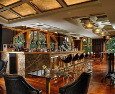 A barman stands behind a marble bar in an upmarket lounge with leather chairs, bottles on display, and large windows letting in natural light.