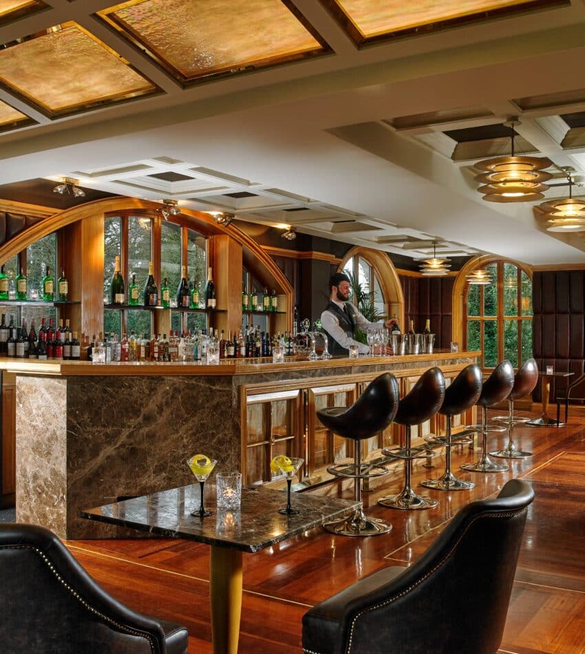 A barman stands behind a marble bar in an upmarket lounge with leather chairs, bottles on display, and large windows letting in natural light.