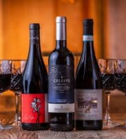 Three bottles of red wine are displayed in front of six filled wine glasses on a wooden table with a warm, blurred background.