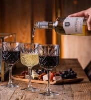 A hand pours white wine into a glass beside two glasses of red wine on a wooden table with cheese, nuts, grapes, and a lit candle.