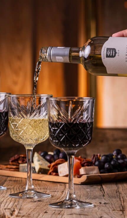 A hand pours white wine into a glass beside two glasses of red wine on a wooden table with cheese, nuts, grapes, and a lit candle.