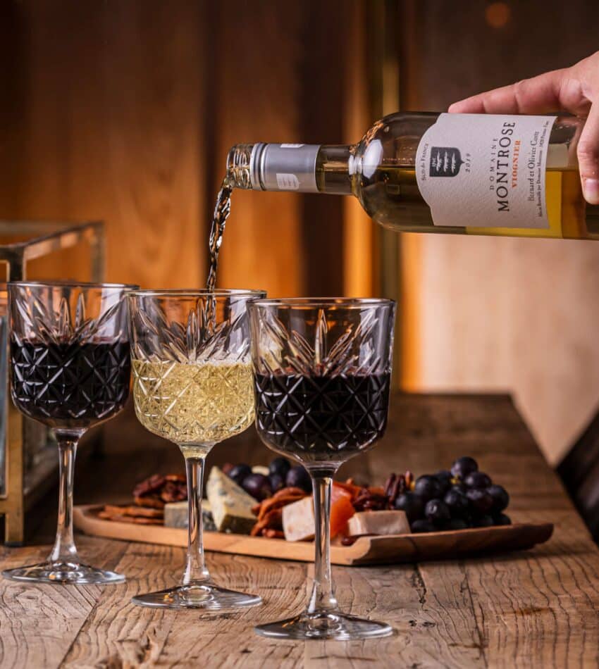 A hand pours white wine into a glass beside two glasses of red wine on a wooden table with cheese, nuts, grapes, and a lit candle.