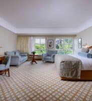 Spacious hotel room with a large bed, seating area with sofa and chairs, wooden desk, TV, and windows overlooking greenery.