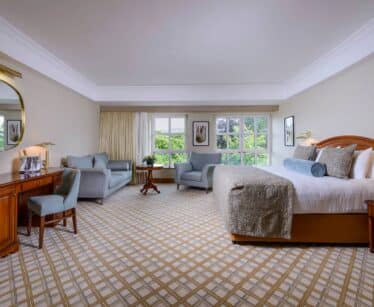 Spacious hotel room with a large bed, seating area with sofa and chairs, wooden desk, TV, and windows overlooking greenery.