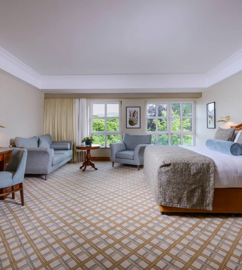 Spacious hotel room with a large bed, seating area with sofa and chairs, wooden desk, TV, and windows overlooking greenery.