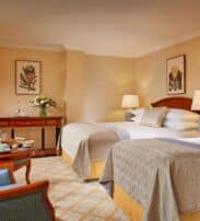Hotel room with two double beds, armchairs around a small table with breakfast, desk with flowers, table lamps, and framed botanical prints on beige walls.