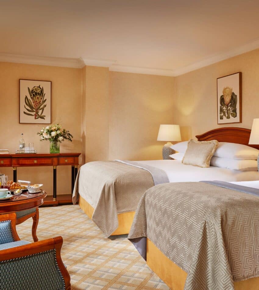 Hotel room with two double beds, armchairs around a small table with breakfast, desk with flowers, table lamps, and framed botanical prints on beige walls.