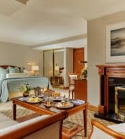 A hotel suite with a bed, seating area, and coffee table set for breakfast. There is a fireplace with a landscape painting above it and a large mirror on the wall.